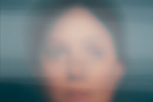 A person's face in a horizontal blurry photograph with white layers over the photograph obscuring the facial features of the person. 